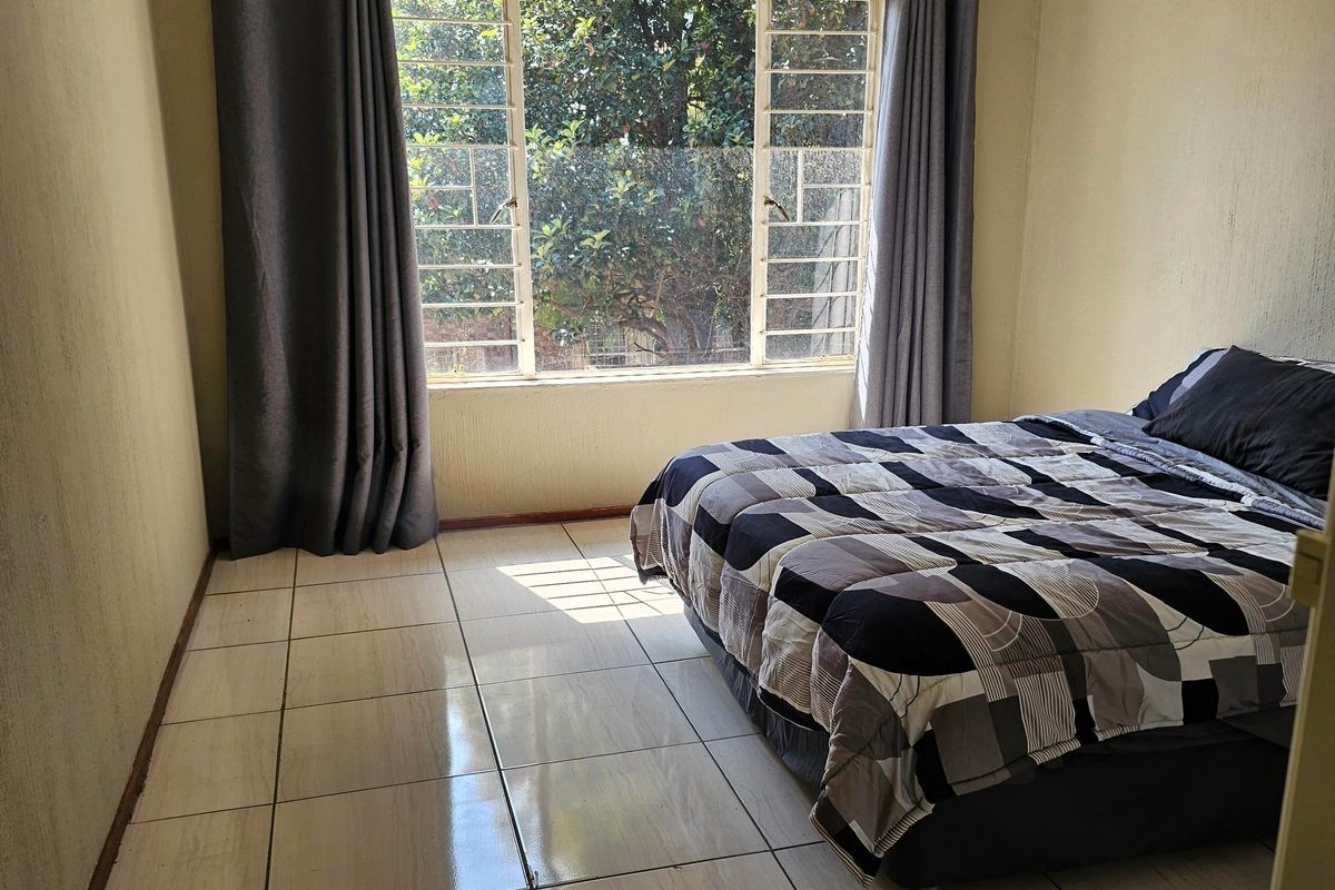 A bedroom featuring tiled flooring and a large window with security bars, providing natural light and a view of the surrounding greenery.