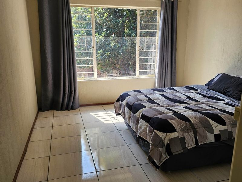 A bedroom featuring tiled flooring and a large window with security bars, providing natural light and a view of the surrounding greenery.