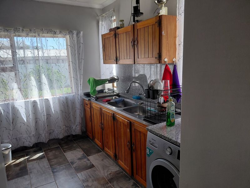 A functional laundry area featuring wooden cabinetry, granite countertops, a double stainless steel sink, and tiled flooring with space for a washing machine.