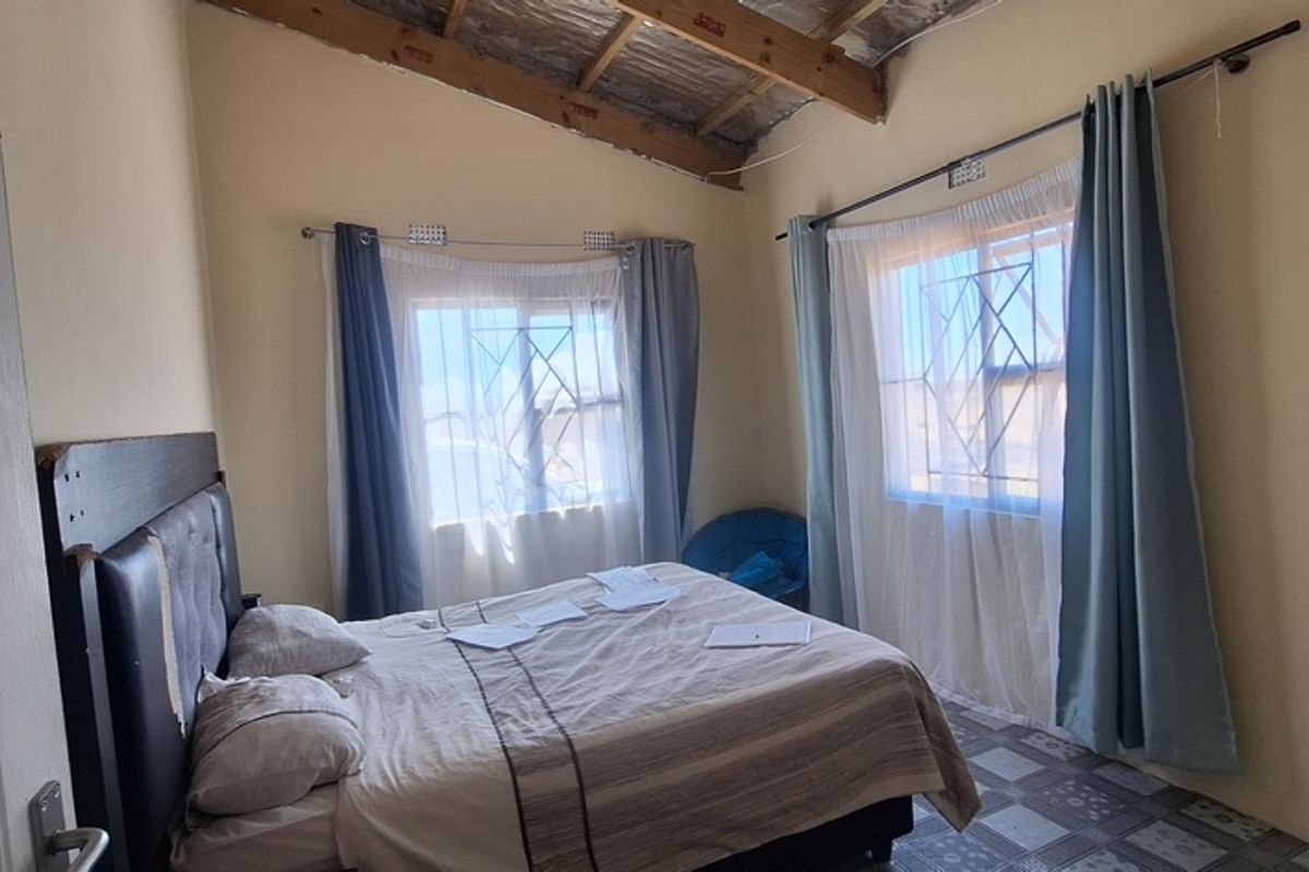 A bedroom featuring exposed timber rafters, security burglar bars on windows, and tiled flooring, currently requiring ceiling completion and general cosmetic updates.