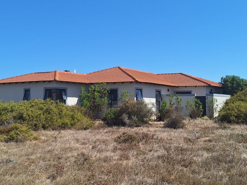 A rear view of a single-story residential house featuring a terracotta tiled roof and stucco exterior walls, surrounded by natural coastal vegetation.