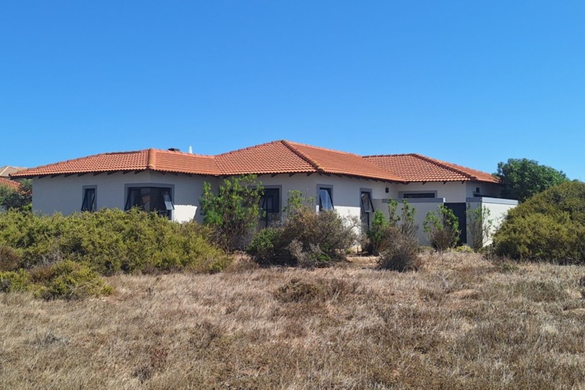 A rear view of a single-story residential house featuring a terracotta tiled roof and stucco exterior walls, surrounded by natural coastal vegetation.