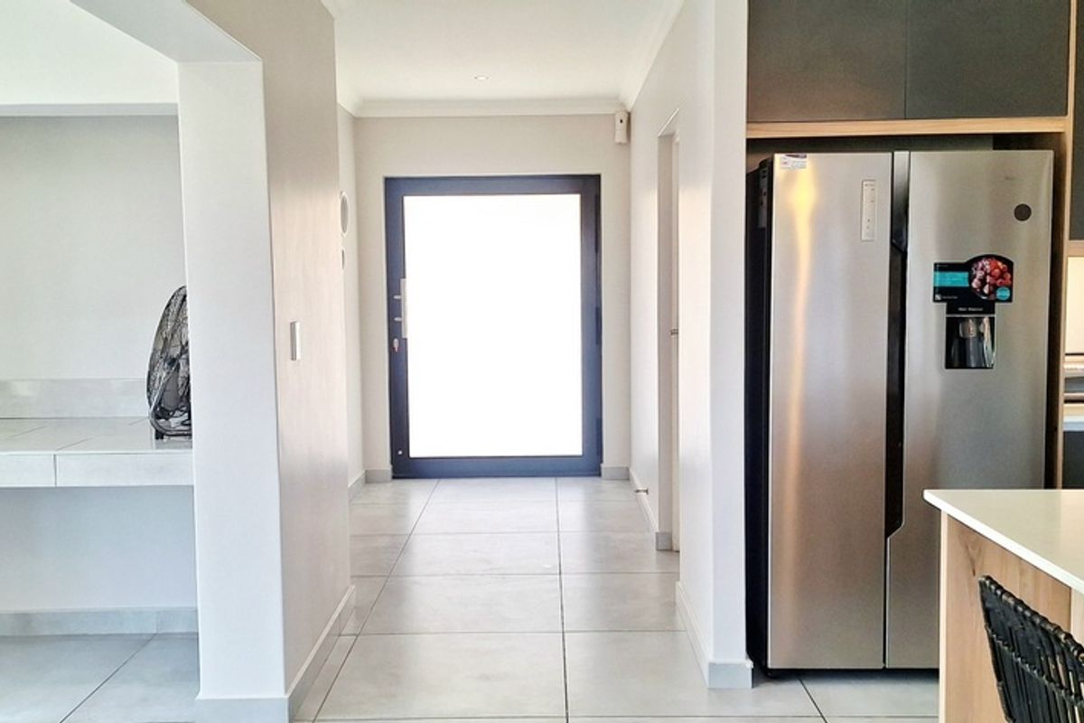 Modern kitchen featuring sleek cabinetry, stone countertops, and tiled flooring, leading into a bright, open-plan living area with a glass door entrance.