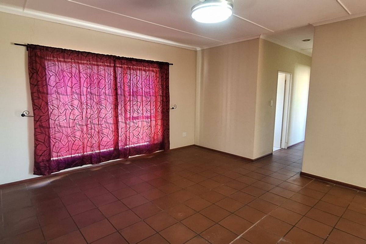 A spacious, empty living area featuring terracotta floor tiles, a large sliding door with curtains, and access to an adjoining hallway.