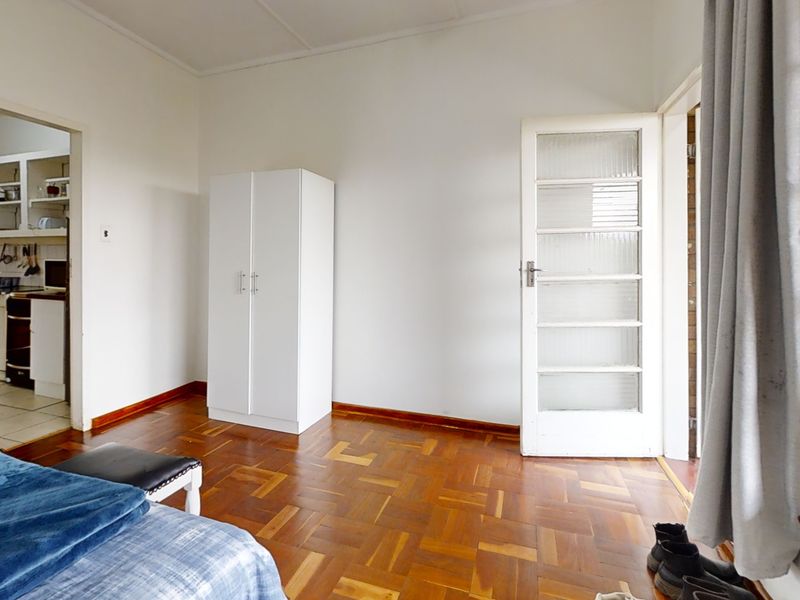 A bedroom featuring classic parquet flooring, a timber-framed glass door leading outside, and an adjacent view into the kitchen area.