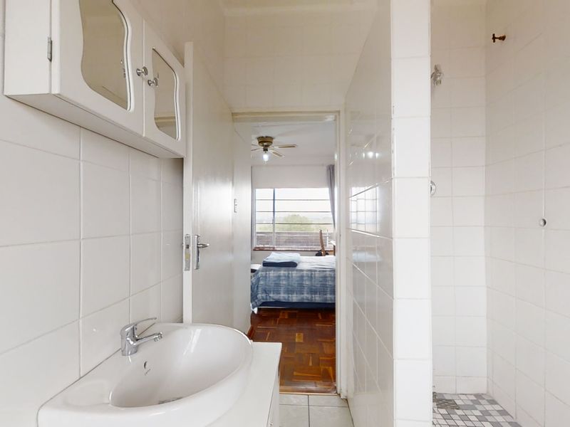 A functional bathroom featuring a wall-mounted vanity, ceramic basin, and a tiled shower enclosure, with a view into the adjacent bedroom.