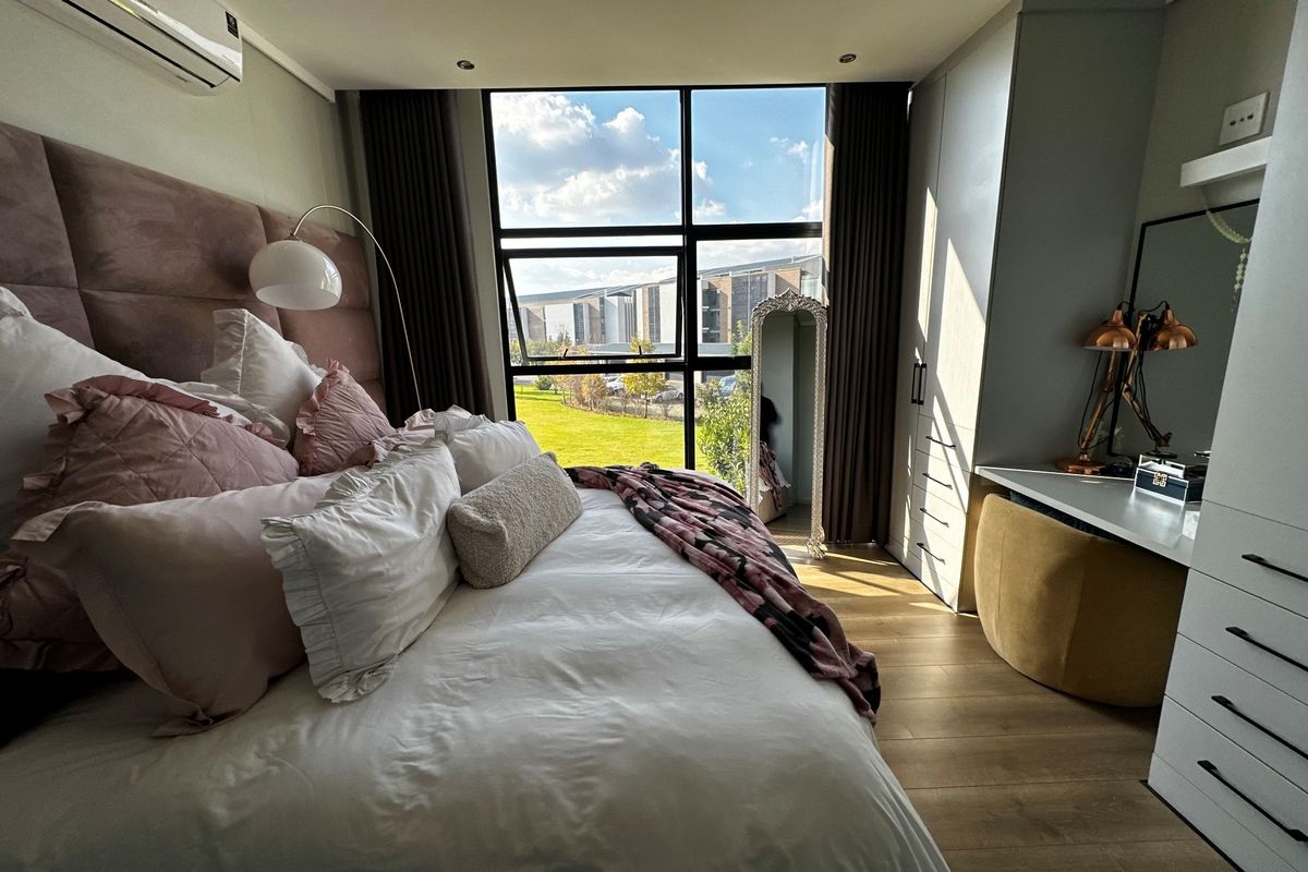 A well-lit bedroom featuring built-in cabinetry, a desk area, large windows with garden views, and a wall-mounted air conditioning unit.