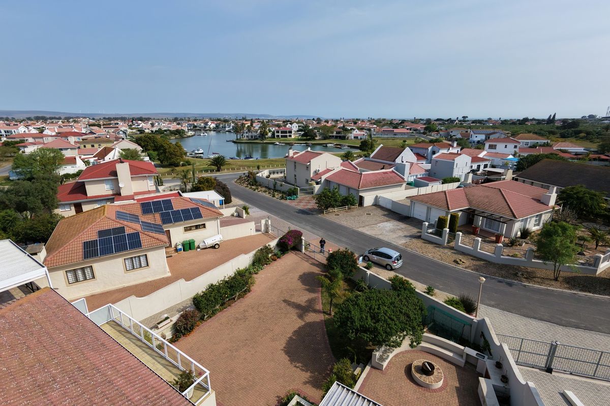 Aerial view of a residential estate featuring paved driveways, solar panels, and a communal fire pit area with surrounding neighborhood and lagoon views.