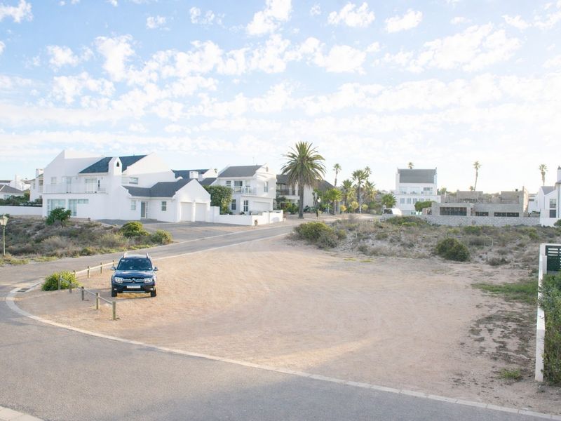 A vacant residential plot featuring a gravel driveway and timber bollards, situated in a coastal neighborhood setting.