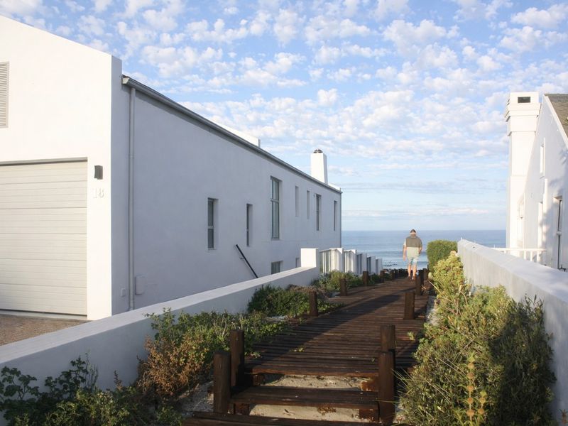 A view of the rear exterior featuring a wooden boardwalk leading towards the ocean, adjacent to a white residential building with a garage.