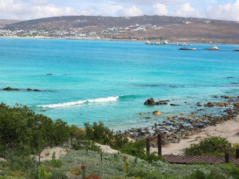 A scenic view of the turquoise ocean, rocky shoreline, and distant coastal hills, featuring a wooden path leading toward the beach.