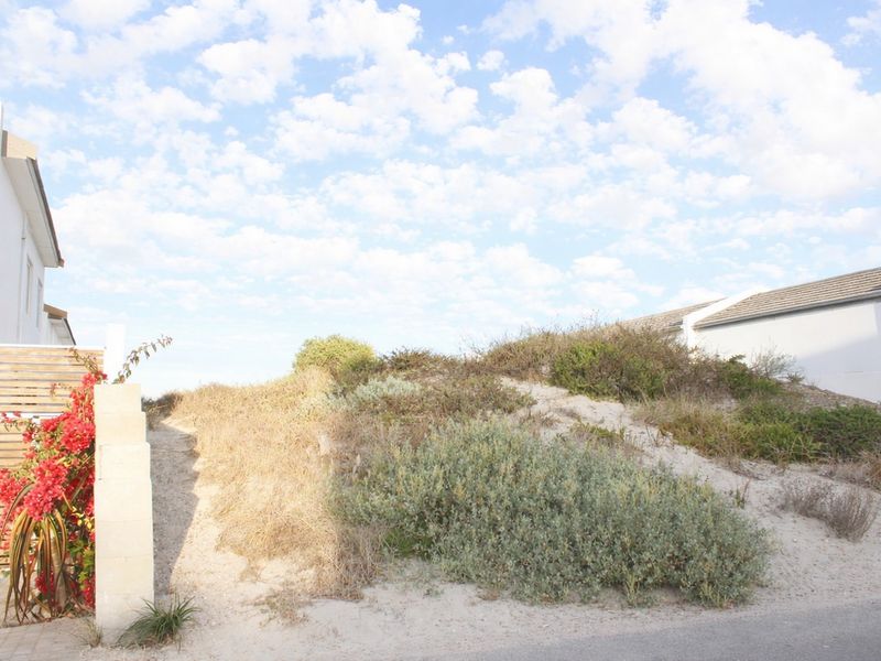 A view of a residential vacant land plot featuring a natural sand dune with coastal vegetation, adjacent to existing residential structures.