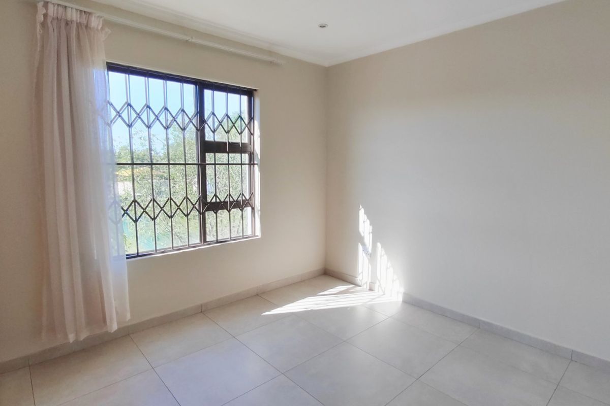 A bright, empty bedroom featuring neutral tiled flooring, built-in cupboards, and a window fitted with security bars.