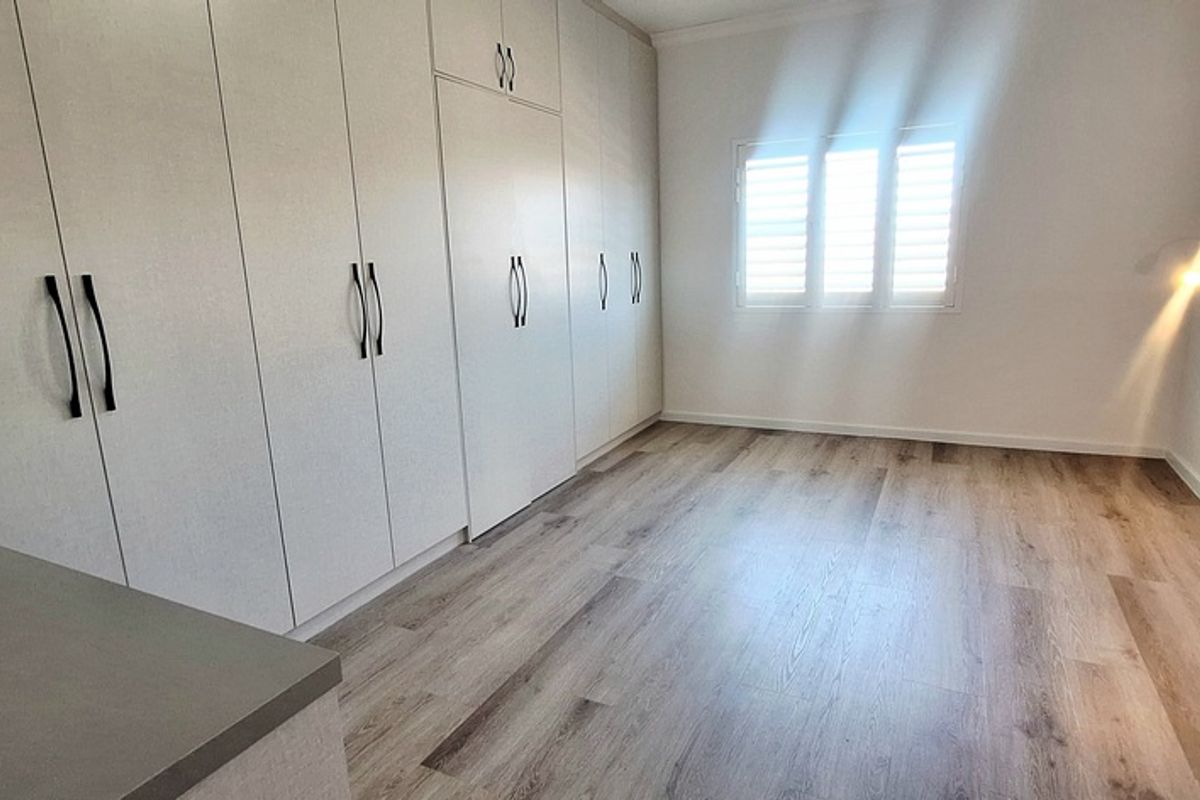A bright, modern bedroom featuring extensive built-in cabinetry, light wood-look flooring, and plantation shutters, offering a clean and functional living space.