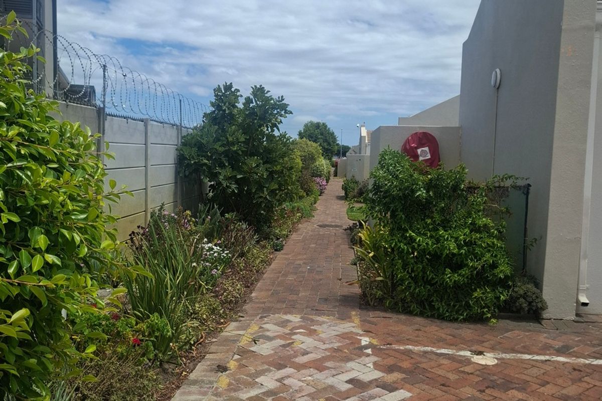 A paved walkway leading through a landscaped garden area between apartment building structures, featuring established greenery and perimeter security fencing.