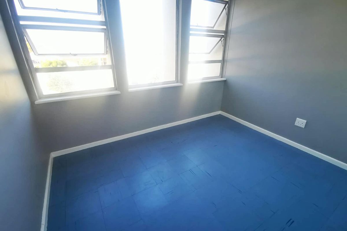 A bright, empty bedroom featuring painted wooden flooring, neutral grey walls, and large top-hung windows providing ample natural light.