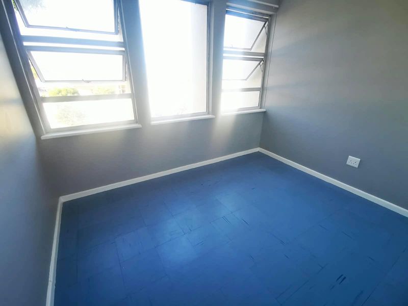 A bright, empty bedroom featuring painted wooden flooring, neutral grey walls, and large top-hung windows providing ample natural light.
