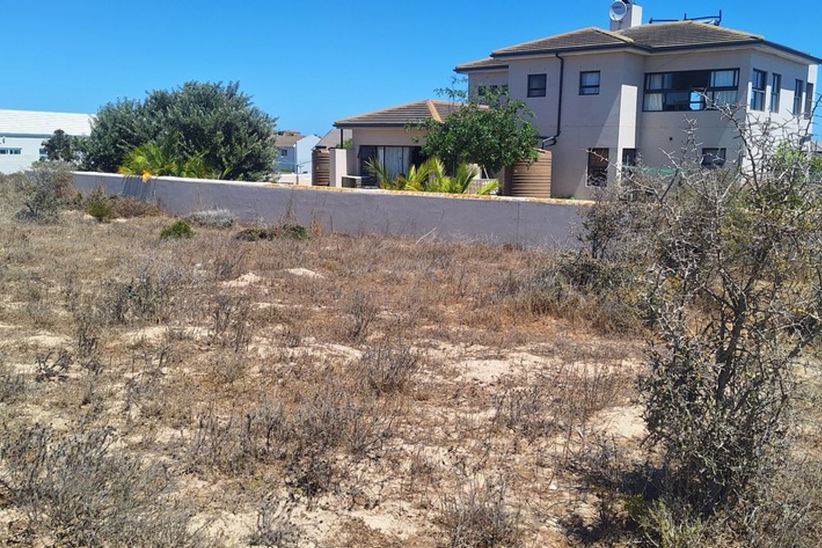 A view of a vacant residential plot featuring natural vegetation, a boundary wall, and adjacent residential properties.