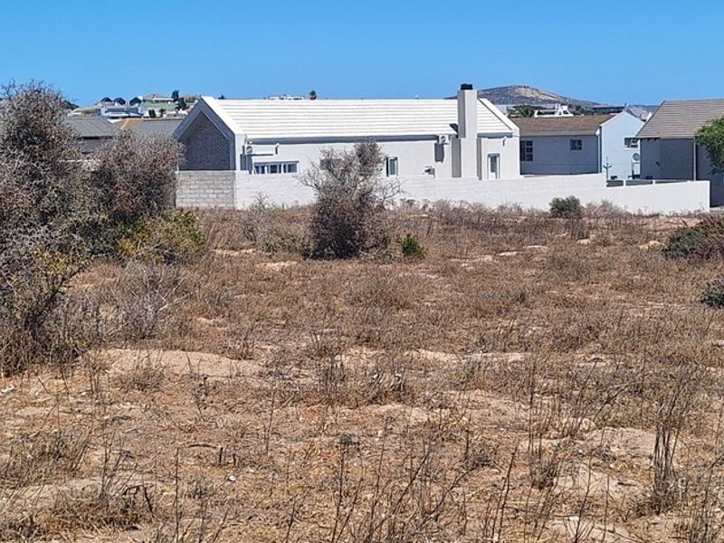 A vacant residential plot featuring natural scrub vegetation with established residential properties visible in the background.
