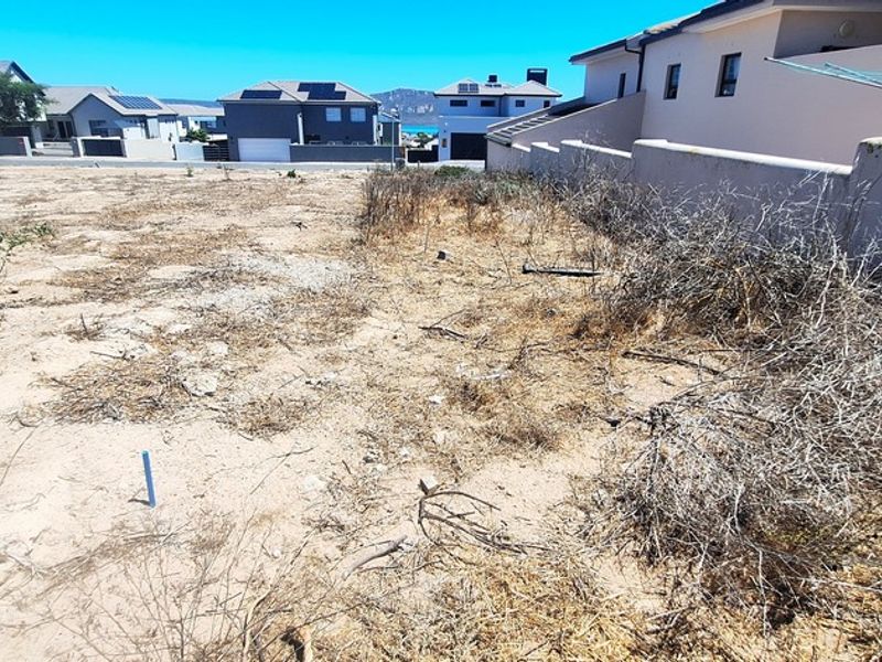 A vacant residential plot featuring sandy terrain and a boundary wall, situated in a developed neighborhood with surrounding residential properties.