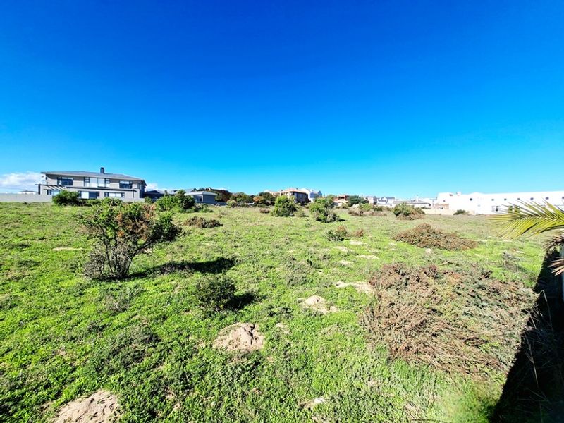 A wide-angle view of a vacant residential plot featuring natural vegetation and surrounding neighborhood homes under a clear blue sky.