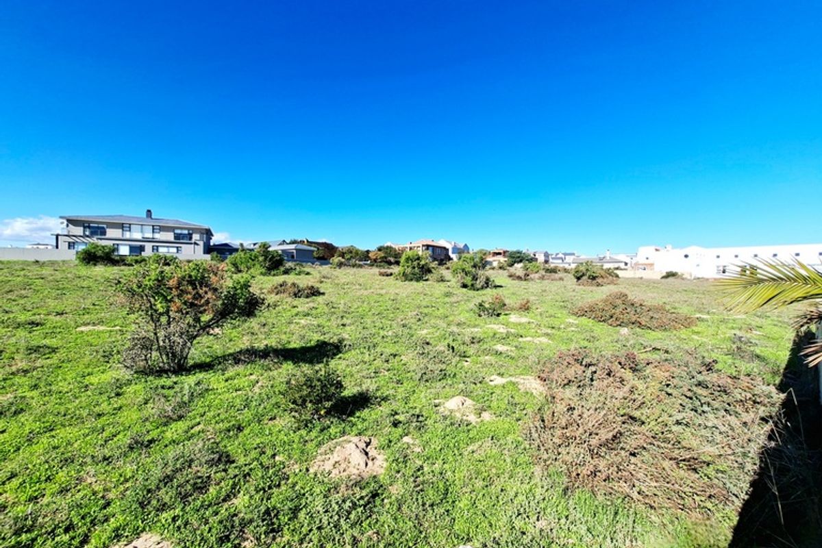 A wide-angle view of a vacant residential plot featuring natural vegetation and surrounding neighborhood homes under a clear blue sky.