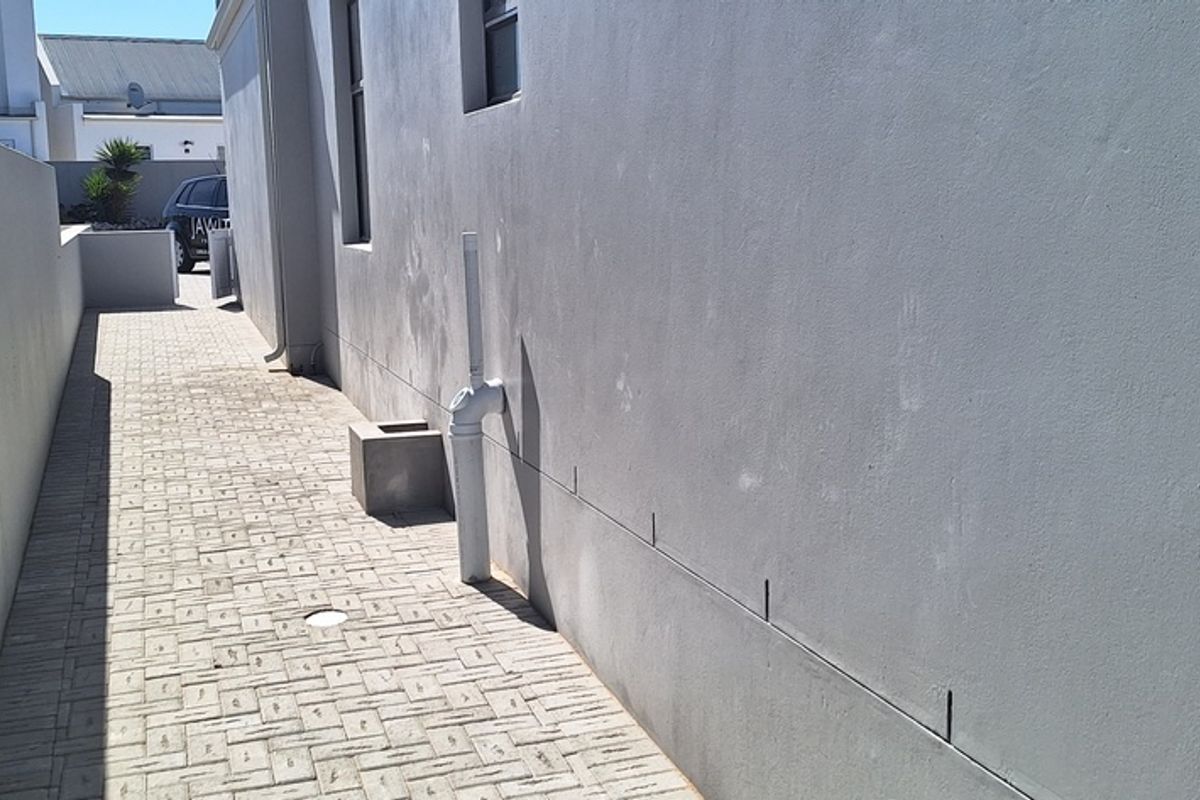 A narrow paved side passage or rear walkway featuring a rendered boundary wall and external plumbing infrastructure.