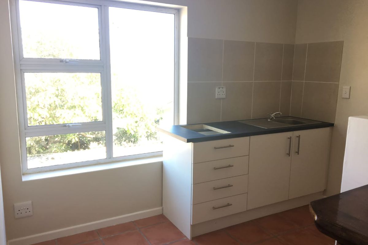 A functional kitchen area featuring built-in cabinetry, a stainless steel sink, tiled splashback, and terracotta flooring, illuminated by a large window.