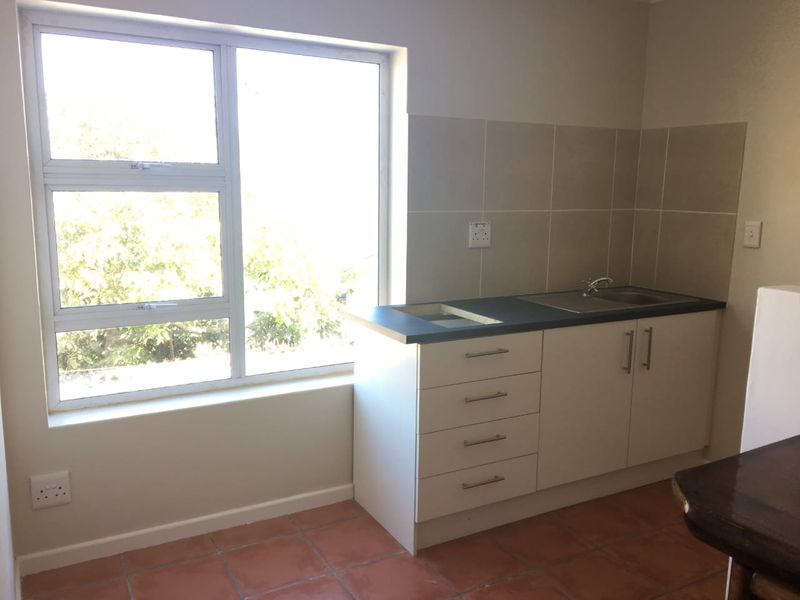 A functional kitchen area featuring built-in cabinetry, a stainless steel sink, tiled splashback, and terracotta flooring, illuminated by a large window.
