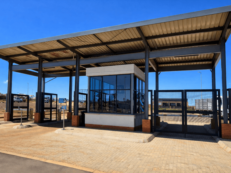 A modern security gatehouse featuring a steel-framed canopy, glass-enclosed guard station, and automated metal access gates.