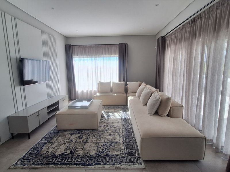 A modern, well-lit living room featuring a custom media wall, large format floor tiles, and expansive windows with floor-to-ceiling curtains.