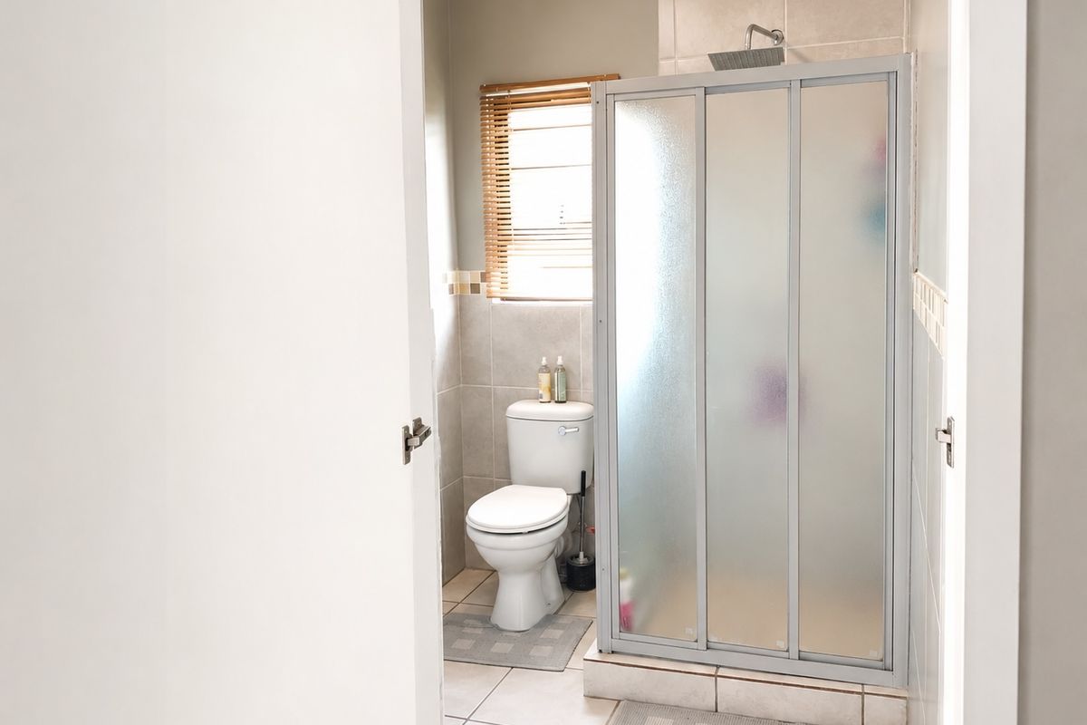 A functional bathroom featuring a tiled shower enclosure with a sliding door, a standard toilet, and a window with fitted blinds.