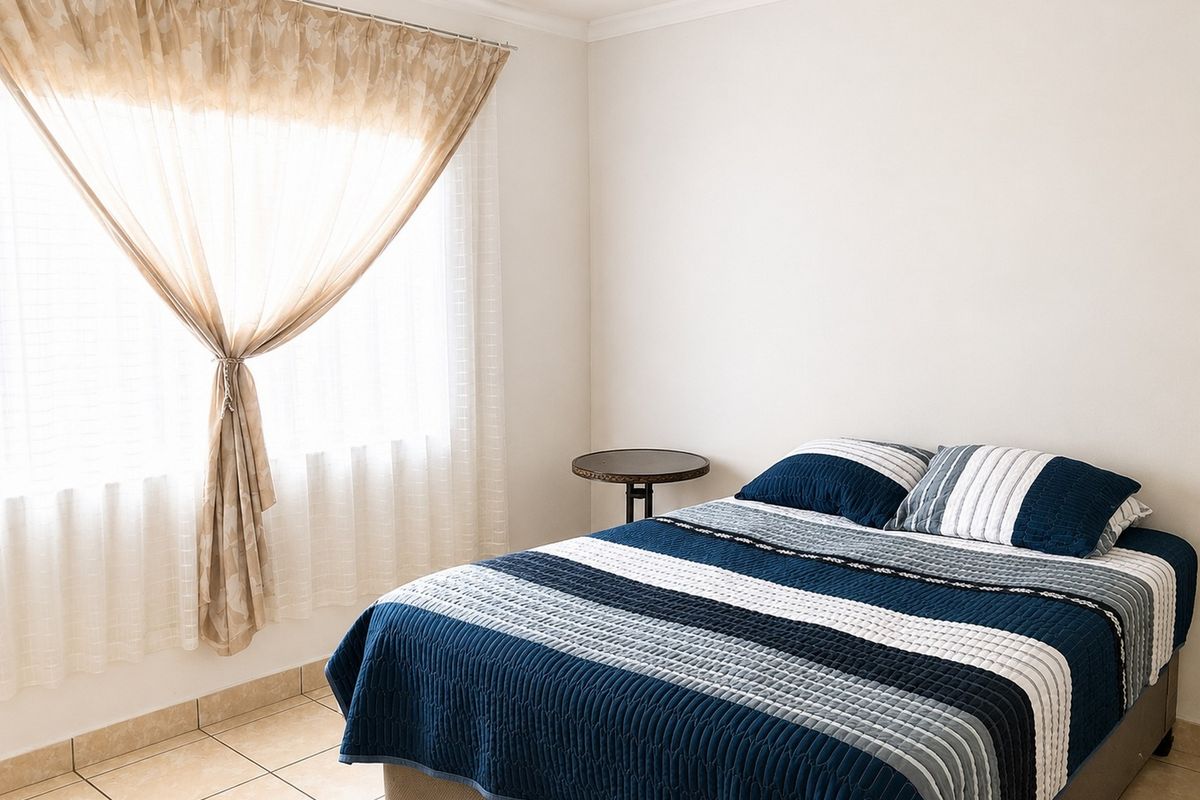 A bright bedroom featuring tiled flooring and a large window with neutral curtains, providing a clean and functional living space.