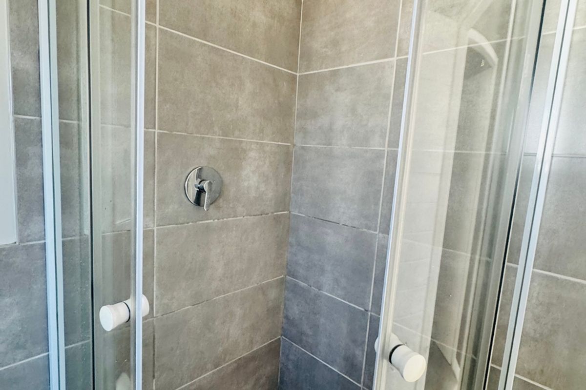 A modern shower enclosure featuring grey wall tiles, a glass door, and a chrome shower head and mixer tap.