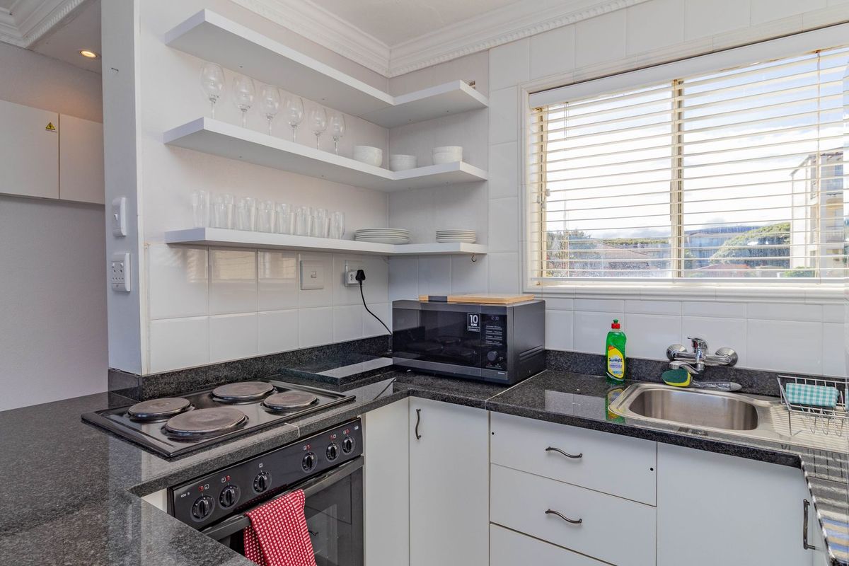 Compact kitchen featuring granite countertops, electric hob, built-in oven, stainless steel sink, and open shelving with a tiled backsplash.