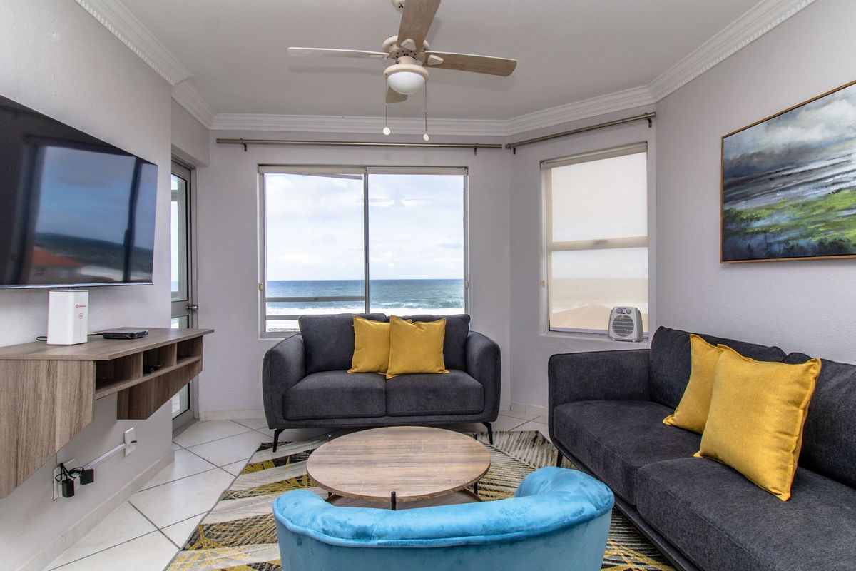 A bright living room featuring tiled floors, a ceiling fan, and large windows offering direct ocean views.