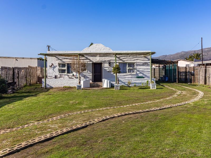 A charming single-story cottage featuring a white brick facade, a covered patio area, and a spacious front lawn with paved driveway tracks.