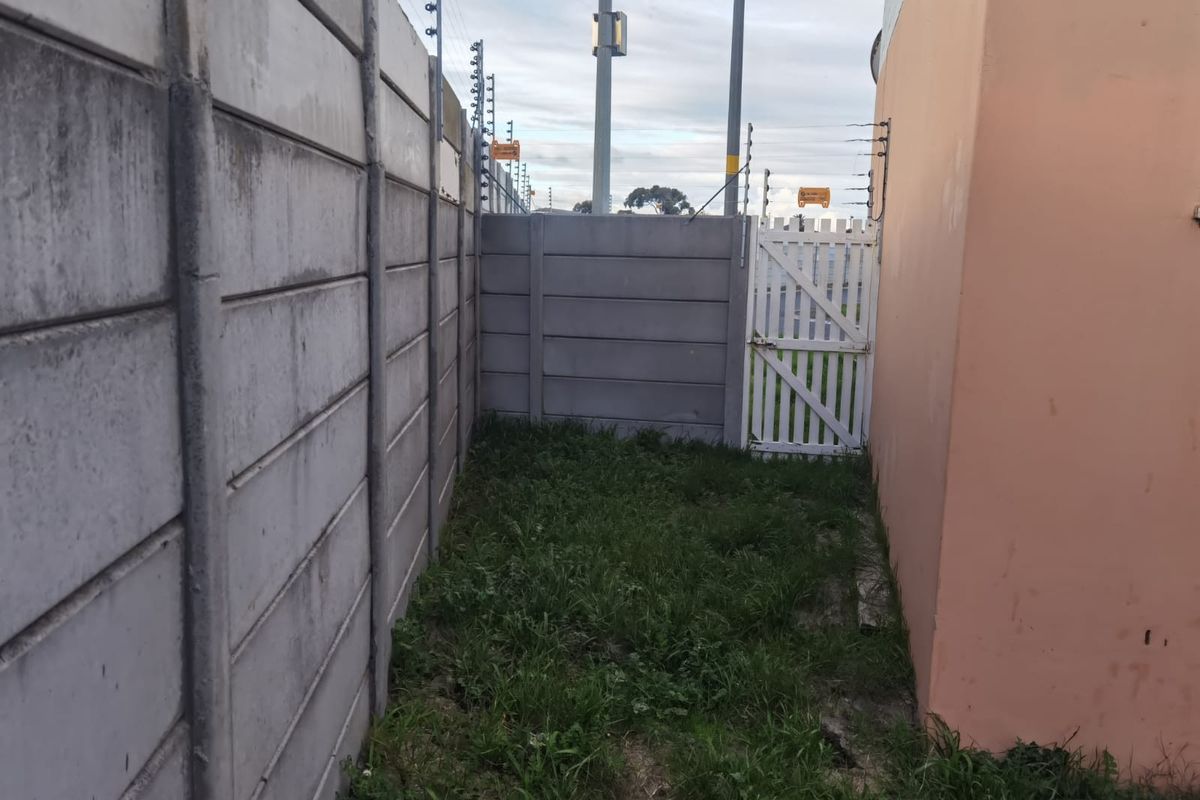 A narrow side passage featuring concrete boundary walls, an electric fence, and a small white pedestrian gate leading to the rear area.