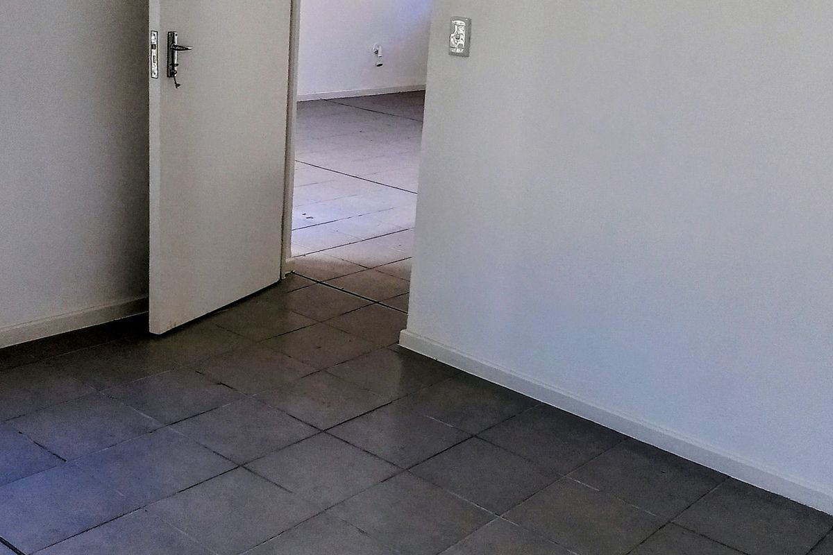An empty bedroom featuring grey tiled flooring, a window with a security gate, and a standard internal door leading to an adjacent room.