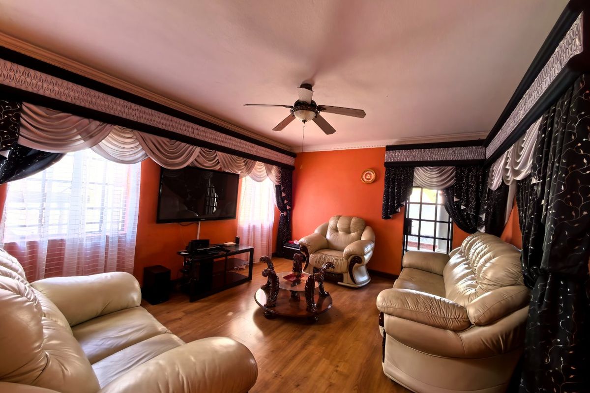 A living room featuring laminate flooring, a ceiling fan, and large windows with decorative valances.