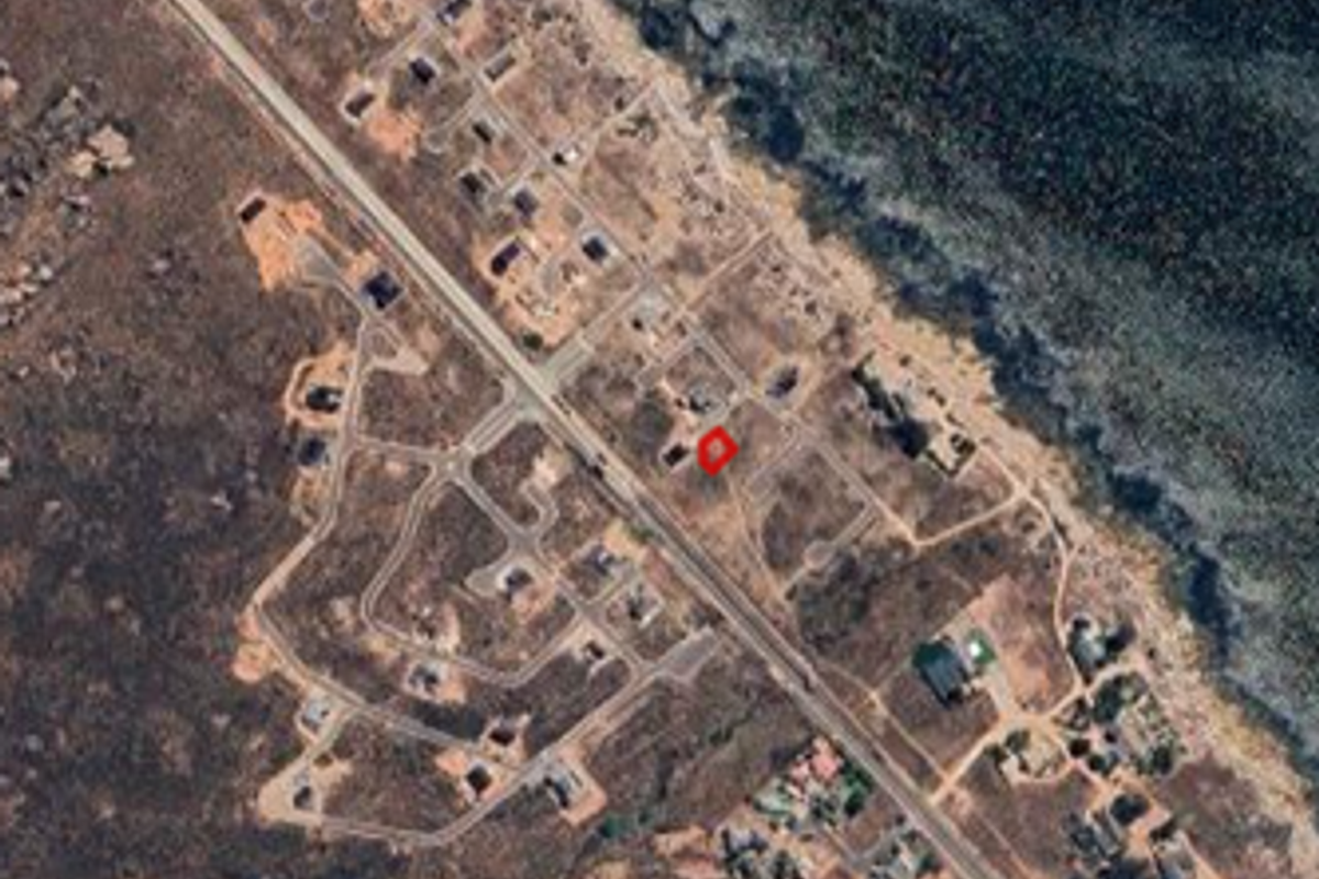 An aerial view of a vacant residential plot within a coastal development, showing surrounding road infrastructure and proximity to the coastline.