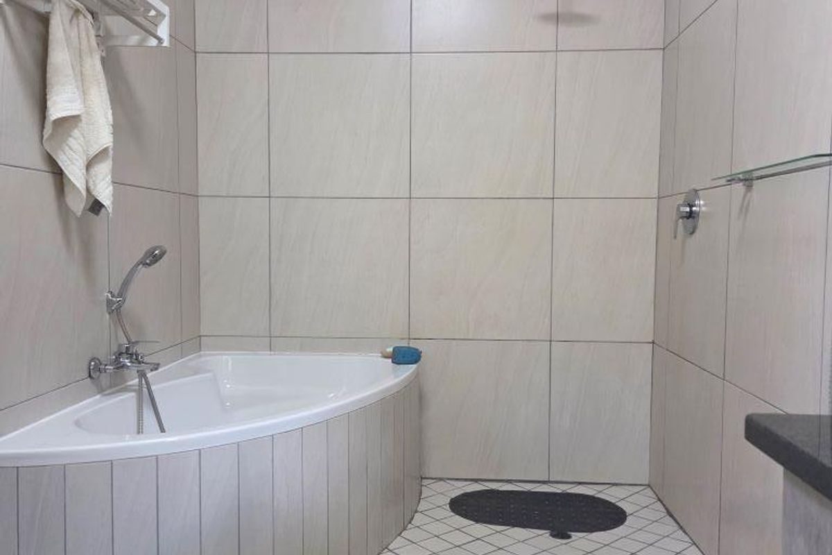 A modern bathroom featuring a corner bathtub, a wall-mounted rainfall shower head, and neutral floor-to-ceiling tiling.