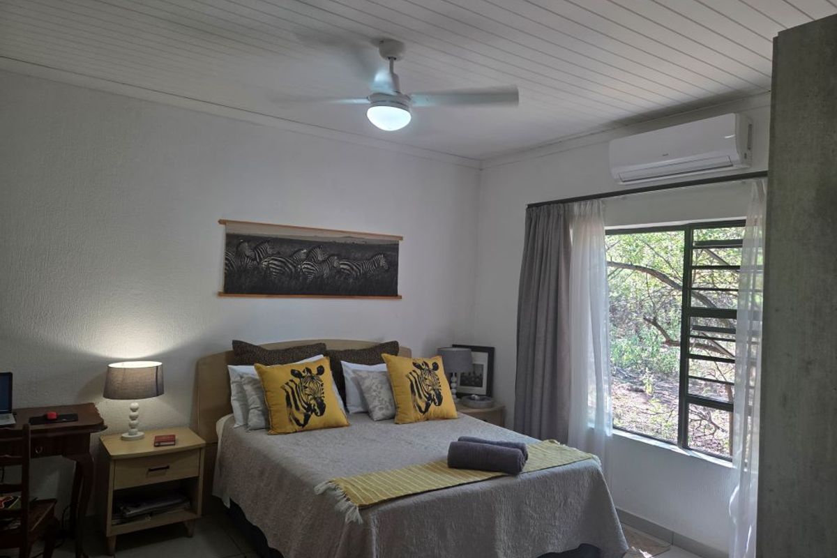 A well-maintained bedroom featuring a ceiling fan, wall-mounted air conditioning unit, and large window with natural light and garden views.