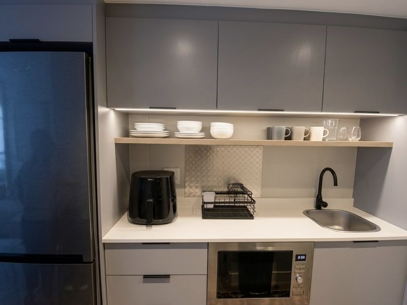 Modern compact kitchen featuring sleek grey cabinetry, stone countertops, integrated microwave, stainless steel sink with matte black faucet, and ambient under-cabinet lighting.