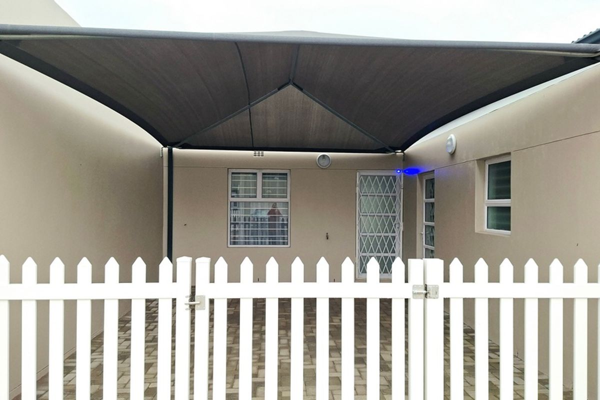 A neat rear exterior area featuring a large shade sail, paved flooring, and a secure white picket fence with gated access.