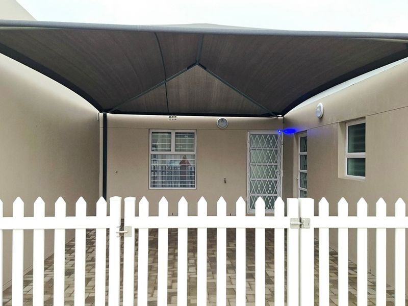 A neat rear exterior area featuring a large shade sail, paved flooring, and a secure white picket fence with gated access.