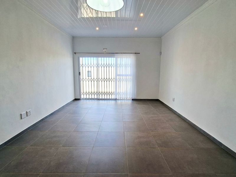 A bright, empty living room featuring large format floor tiles, a tongue and groove ceiling, and a sliding glass door with a security gate.