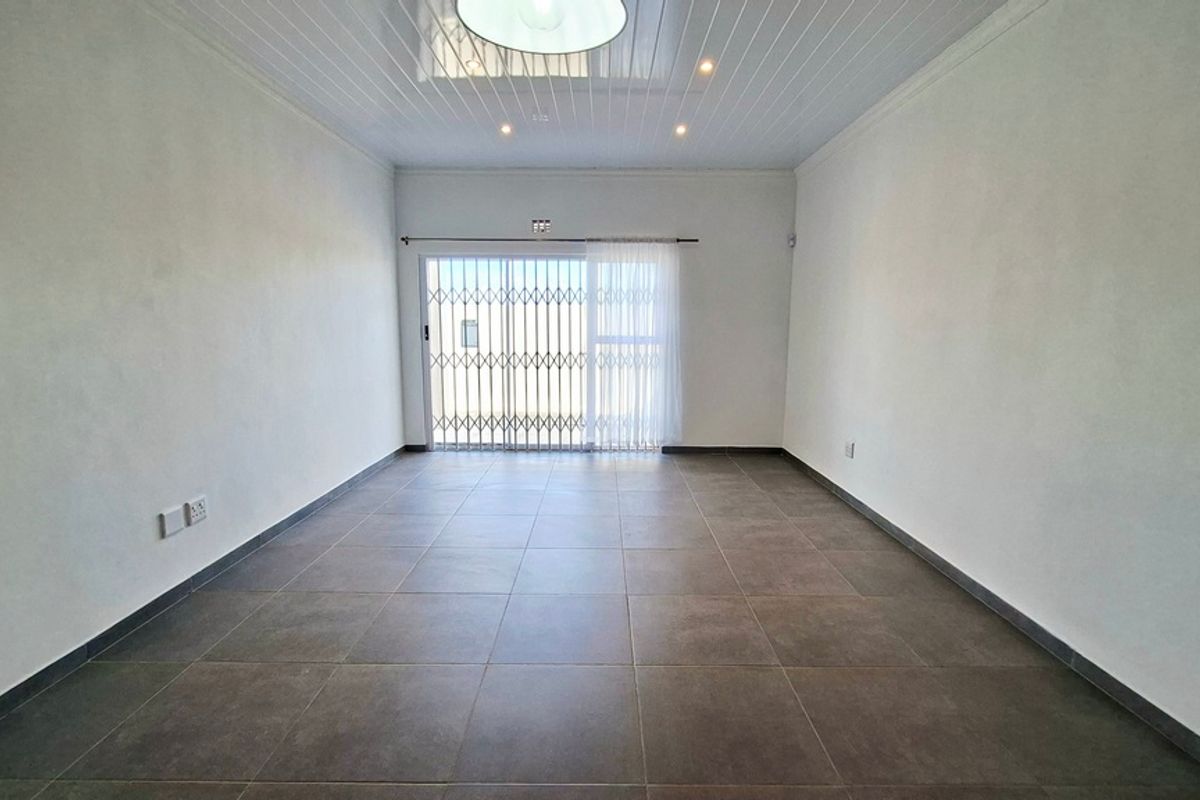 A bright, empty living room featuring large format floor tiles, a tongue and groove ceiling, and a sliding glass door with a security gate.