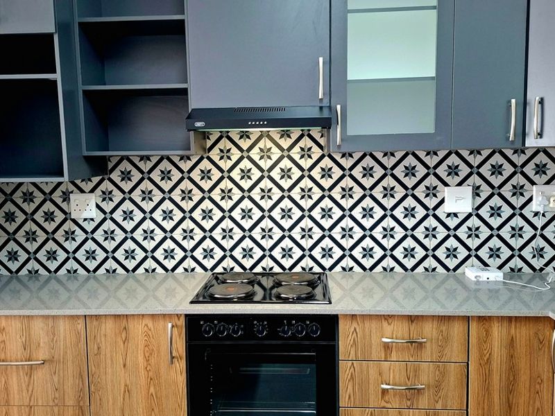 Modern kitchen featuring wood-grain lower cabinetry, dark upper cabinets, stone countertops, and a decorative patterned tile backsplash with integrated oven and hob.