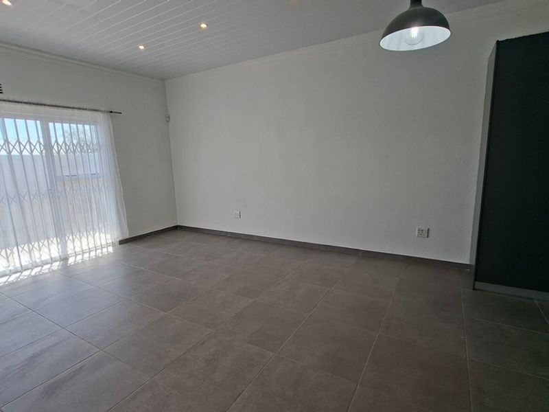 A bright, empty living room featuring large format floor tiles, a sliding door with security gate, and a built-in storage cupboard.